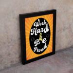 Work Hard Be Proud office wall art with white text on a black effect background gradient of orange and yellow, framed in black on the wall. Perfect for home office decor.