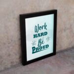 Work Hard Be Proud office wall art featuring green text with decorative elements on a white background, framed in black on the wall. Perfect for home offices.