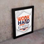 Inspirational quote wall art featuring Work Hard Be Proud with orange and navy blue text on a light gray and light green background and black frame on the wall. Perfect for home offices.