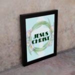 Infidu Jesus Christ Bible Wall Art with JESUS CHRIST text in a mint blue background surrounded by a floral wreath. displayed on the wall in a black frame