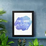 Bible wall art with The Lord Is My Rock, My Fortress, and My Deliverer in white stencil-style text on a blue watercolor background. displayed on the wall in a black frame