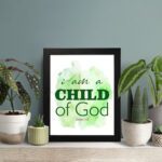 I Am A Child Of God wall art in elegant fonts with a green and black color scheme on a soft green watercolor background. displayed on the table in a black frame