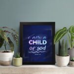 Infidu I Am A Child Of God wall art featuring soft glowing text in pink and blue on a dark blue background. displayed on the table in a black frame