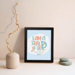 I Am a Child of God Christian Wall Art in bright orange and turquoise with a pastel blue background, perfect for home decor. displayed on the wall in a black frame