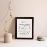 Infidu wall art with Proverbs 4:7 text surrounded by green leaves on a beige background. Elegant and minimal design. displayed on the wall in a black frame