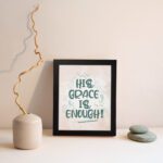 Christian wall art featuring the phrase His Grace Is Enough in teal lettering on a soft pink background with decorative swirls. displayed on the wall in a black frame