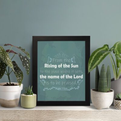 Wall art displaying Psalm 113:3, featuring white text on a dark green background with butterfly and leaf designs. displayed on the table in a black frame