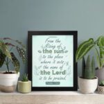 Wall art displaying Psalm 113:3 with elegant cursive font and leafy motifs on a mint green background. displayed on the table in a black frame