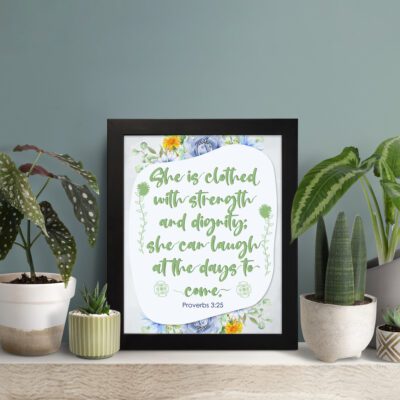 Christian wall art featuring Proverbs 31:25, with elegant script and floral accents in light green and gray background. displayed on the table in a black frame