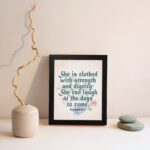 Wall art featuring Proverbs 31:25 with the quote in dark green text and floral decorations on a cream background. displayed on the wall in a black frame