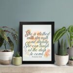 Christian wall art featuring the quote She is clothed with strength and dignity; she can laugh at the days to come from Proverbs 31:25, with an elegant design and pastel colors. displayed on the table in a black frame