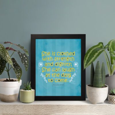 Christian wall art with Proverbs 31:25 verse in bold yellow text on a dark blue background with floral accents displayed on the table in a black frame