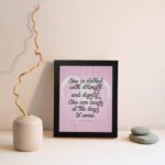 Proverbs 31:25 Christian wall art with She is clothed with strength and dignity on a soft pink background in an elegant script, framed. displayed on the wall in a black frame