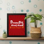 Infidu Dream Big Work Hard Make It Happen wall art with a red background white and yellow text and a black frame on the table. Inspiring decor for home offices and workspaces.