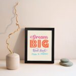 Infidu Dream Big Work Hard Make It Happen wall art with a light pink background and vibrant pink, orange, violet, and sky blue text and a black frame displayed on the wall. Inspiring decor for home offices and workspaces.