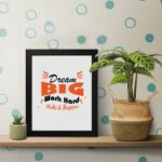 Infidu Dream Big Work Hard Make It Happen wall art with a gray background black and orange text and a black frame on the table. Inspiring decor for home offices and workspaces.