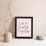 Infidu Take A Small Step Everyday wall art with a gray and light pink background black and red text and a black frame on the wall. Inspiring decor for any workspace.