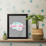 Infidu Take A Small Step Everyday wall art with a gray background and vibrant green, yellow, and pink text and black frame on the table. Inspiring decor for any workspace.