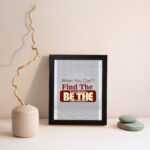 Infidu When You Can't Find The Sunshine Be The Sunshine wall art with a gray and white background and red text and black frame displayed on the wall Inspiring decor for any workspace.