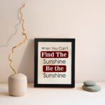 Infidu motivational wall art featuring the quote When You Can't Find the Sunshine Be the Sunshine on a white background with red text and a black frame displayed on the wall. perfect for office decor.