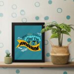 Infidu When You Can't Find the Sunshine, Be the Sunshine Office Wall Art featuring black, yellow, and blue text on a light blue background with decorative elements, framed in black on the table. Perfect for inspirational quote wall art, office wall art, and home decor.