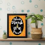 Work Hard Be Proud office wall art with white text on a black effect background gradient of orange and yellow, framed in black on the table. Perfect for home office decor.