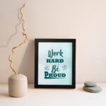 Work Hard Be Proud office wall art featuring green text with decorative elements on a white background, framed in black displayed on the wall. Perfect for home offices.