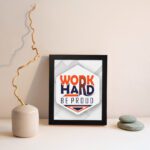 Inspirational quote wall art featuring Work Hard Be Proud with orange and navy blue text on a light gray and light green background and black frame displayed on the wall. Perfect for home offices.