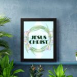 Infidu Jesus Christ Bible Wall Art with JESUS CHRIST text in a mint blue background surrounded by a floral wreath. displayed on the wall in a black frame