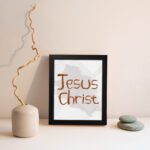 Jesus Christ Bible wall art with hand-drawn brown lettering and a minimalist white-gray watercolor background in a black frame. displayed on the wall in a black frame