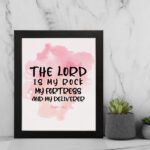 Bible wall art featuring The Lord Is My Rock, My Fortress, and My Deliverer in black handwritten-style lettering on a soft pink watercolor background. displayed on the wall in a black frame