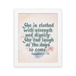 Wall art featuring Proverbs 31:25 with the quote in dark green text and floral decorations on a cream background. hung on the wall in a white frame