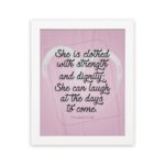 Proverbs 31:25 Christian wall art with She is clothed with strength and dignity on a soft pink background in an elegant script, framed. hung on the wall in a white frame