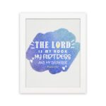 Bible wall art with The Lord Is My Rock, My Fortress, and My Deliverer in white stencil-style text on a blue watercolor background. hung on the wall in a white frame