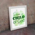 I Am A Child Of God wall art in elegant fonts with a green and black color scheme on a soft green watercolor background. displayed on the wall in a white frame