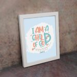 I Am a Child of God Christian Wall Art in bright orange and turquoise with a pastel blue background, perfect for home decor. displayed on the wall in a white frame