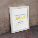 Christian wall art featuring the phrase Your Right Hand Upholds Me in playful fonts with a yellow cursive style and decorative waves. displayed on the wall in a white frame