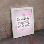 Christian wall art displaying We Walk By Faith Not By Sight with floral elements on a pale pink background. displayed on the wall in a white frame