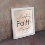 Christian wall art displaying the quote We Walk By Faith Not By Sight in elegant fonts on a soft beige-pink background. displayed on the wall in a white frame