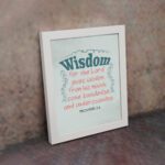 Wisdom Proverbs 2:6 Christian wall art with teal and red text on a mint green background in a black frame. displayed on the wall in a white frame