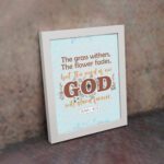 The Grass Withers Christian wall art with the verse Isaiah 40:8 in a modern font, featuring floral illustrations and a light blue background. displayed the wall in a white frame