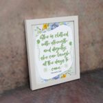 Christian wall art featuring Proverbs 31:25, with elegant script and floral accents in light green and gray background. displayed on the wall in a white frame