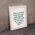 Wall art featuring Proverbs 31:25 with the quote in dark green text and floral decorations on a cream background. displayed on the wall in a white frame