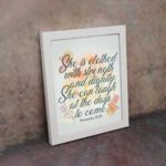 Christian wall art featuring the quote She is clothed with strength and dignity; she can laugh at the days to come from Proverbs 31:25, with an elegant design and pastel colors. displayed on the wall in a white frame