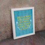 Christian wall art with Proverbs 31:25 verse in bold yellow text on a dark blue background with floral accents displayed on the wall in a white frame