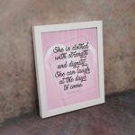 Proverbs 31:25 Christian wall art with She is clothed with strength and dignity on a soft pink background in an elegant script, framed. displayed on the wall in a white frame