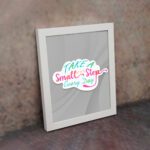 Infidu Take A Small Step Everyday wall art with a gray background and vibrant green, yellow, and pink text and white frame on the wall. Inspiring decor for any workspace.