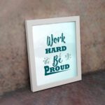 Work Hard Be Proud office wall art featuring green text with decorative elements on a white background, framed in white on the wall. Perfect for home offices.