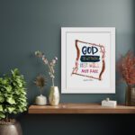 Wall art with the text 'God Is Within Her, She Will Not Fall' in colorful fonts, surrounded by pink blossoms and a wooden frame. displayed on the wall in a white frame