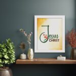 Jesus Christ text in green script with red floral accents on a glowing yellow-orange background, framed in black. displayed on the wall in a white frame